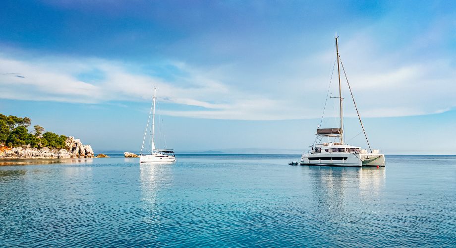 A catamaran and a monohull sailing in the blue ocean. Compare catamarans vs monohulls to find the best fit for your sailing style. Learn about cost, speed, stability, and more.