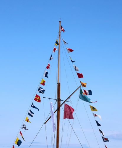 A tall sailboat mast is adorned with a string of vibrant nautical signal yacht flags fluttering in the breeze beneath a clear blue sky.