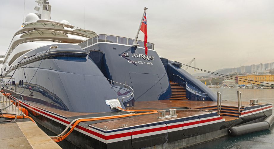 The luxury vessel Al Mirqab, registered in George Town, Cayman Islands, is docked at a marina with a red ensign flying at its stern.