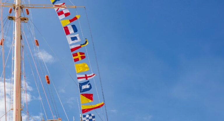 A series of colorful maritime signal yacht flags are strung vertically from a ship’s mast against a clear blue sky. Learn how to choose your yacht flag, understand the flag's meaning, and explore the best boat flags for legal and cultural purposes.