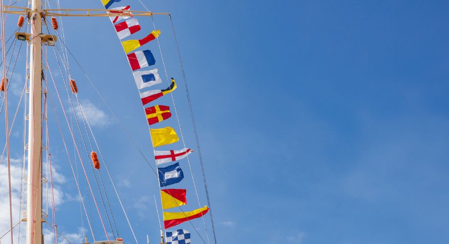 A series of colorful maritime signal yacht flags are strung vertically from a ship’s mast against a clear blue sky. Learn how to choose your yacht flag, understand the flag's meaning, and explore the best boat flags for legal and cultural purposes.