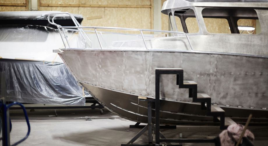 A metal boat hull is under construction in a workshop, with another partially covered vessel in the background.