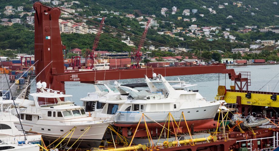 Several vessels are securely fastened on the deck of a cargo ship at a busy port, with a hillside town and shipping containers visible in the background.