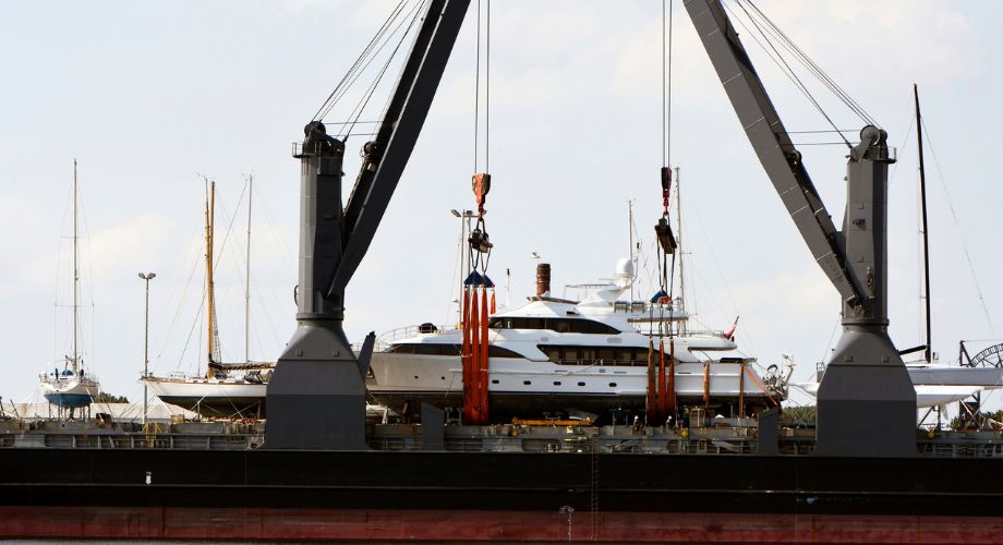 A large luxury yacht is being lifted by heavy-duty cranes onto a cargo ship at a port.