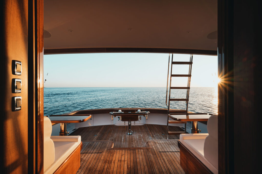 A serene view from inside a wellness superyacht looking out onto calm ocean waters at sunset, with polished wooden decking, cushioned seating, and a ladder leading upwards.