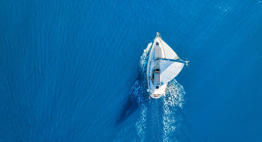A solitary sailboat glides gracefully through calm, crystal-blue waters, captured from above. Its white sails contrast beautifully with the deep azure sea, evoking a sense of serenity, freedom, and sustainable travel.