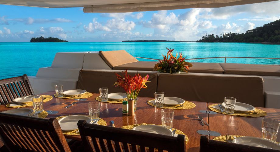 An elegant dining setup on the deck of an eco-friendly yacht overlooks stunning turquoise waters and lush islands, creating a perfect tropical ambiance for an alfresco meal.