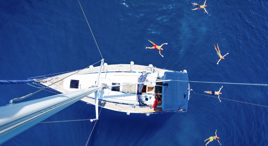 A group of people float joyfully in crystal-clear blue water around a sailboat, captured from above in a stunning aerial view of a perfect summer escape.