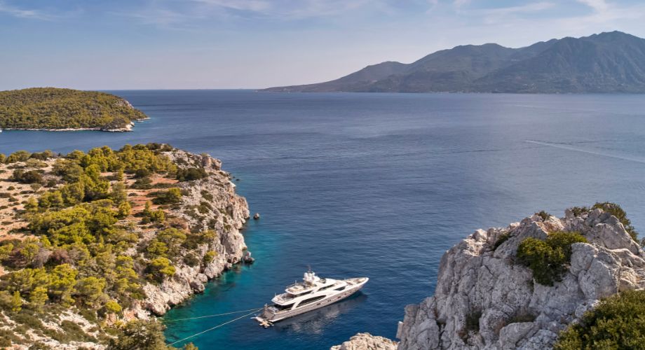 A luxurious yacht is anchored in a secluded turquoise cove surrounded by rocky cliffs and lush greenery, with expansive views of a calm, mountainous coastline.