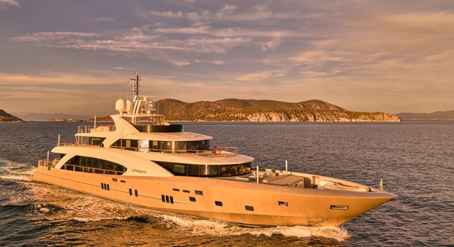 The 50-meter COUACH motor yacht La Pellegrina 1 glides elegantly through calm waters at golden hour, with a scenic coastline and dramatic cliffs in the background.