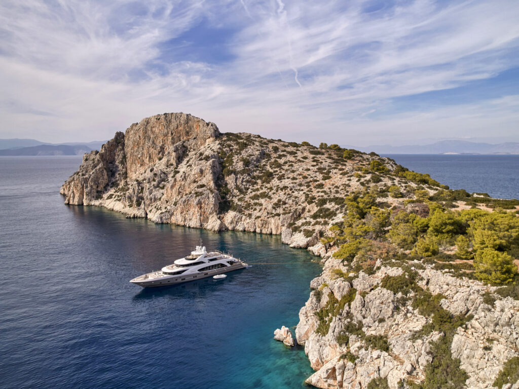 A luxurious yacht is anchored in a secluded turquoise cove surrounded by rocky cliffs and lush greenery, with expansive views of a calm, mountainous coastline.