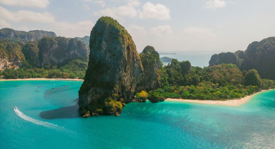 A stunning tropical landscape featuring turquoise waters, lush greenery, white sandy beaches, and a dramatic limestone cliff rising from the sea, with boats dotting the shoreline.