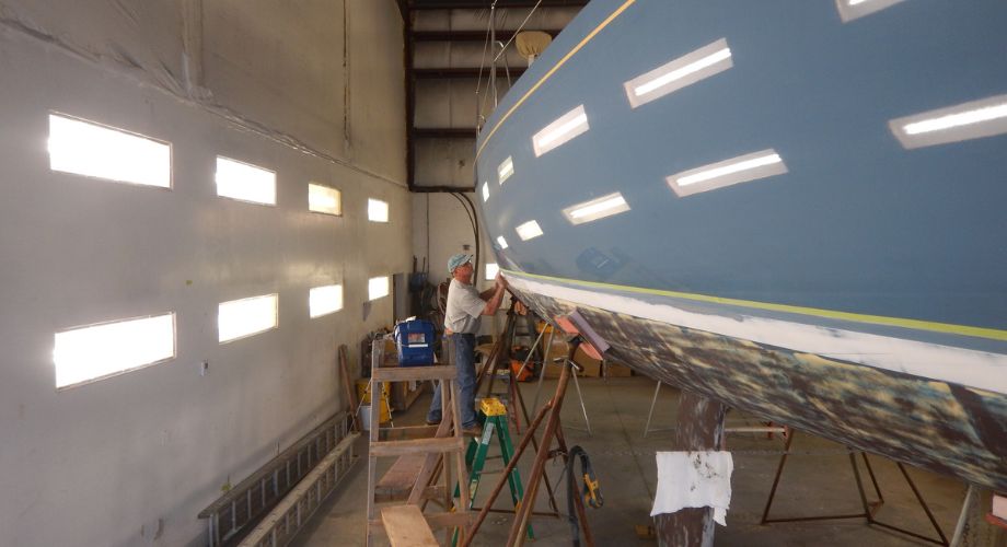 A man is performing maintenance on the hull of a large boat inside a well-lit workshop, with scaffolding, tools, and safety equipment surrounding the vessel.