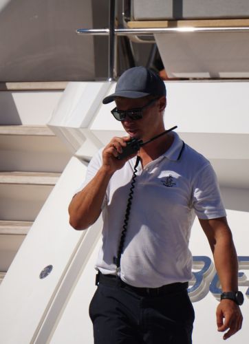 A yacht crew man in a white polo shirt and dark cap is speaking into a radio while standing on the deck of a luxury yacht. Owning a superyacht is more than the purchase price. Discover hidden costs from maintenance to crew and fuel in this clear cost breakdown.