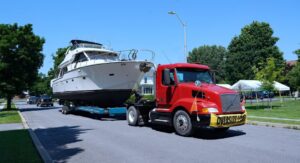 A red semi-truck is transporting a large yacht on a trailer through a residential neighborhood with an "OVERSIZE LOAD" sign, showcasing a yacht delivery in progress. Learn how to prepare for yacht delivery, discover top yacht delivery services, and understand key shipping options for your yacht.