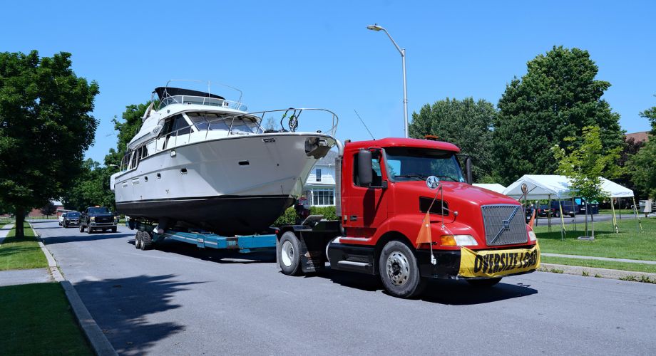 A red semi-truck is transporting a large yacht on a trailer through a residential neighborhood with an "OVERSIZE LOAD" sign, showcasing a yacht delivery in progress. Learn how to prepare for yacht delivery, discover top yacht delivery services, and understand key shipping options for your yacht.