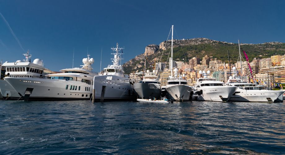 A stunning lineup of superyachts under various yacht registrations anchored along a glamorous marina, with a backdrop of a hillside city and a deep blue sky.