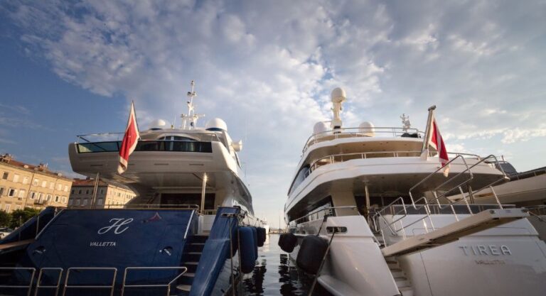 Two luxury yachts with Valletta yacht registration are docked side by side at a marina under a partly cloudy sky.