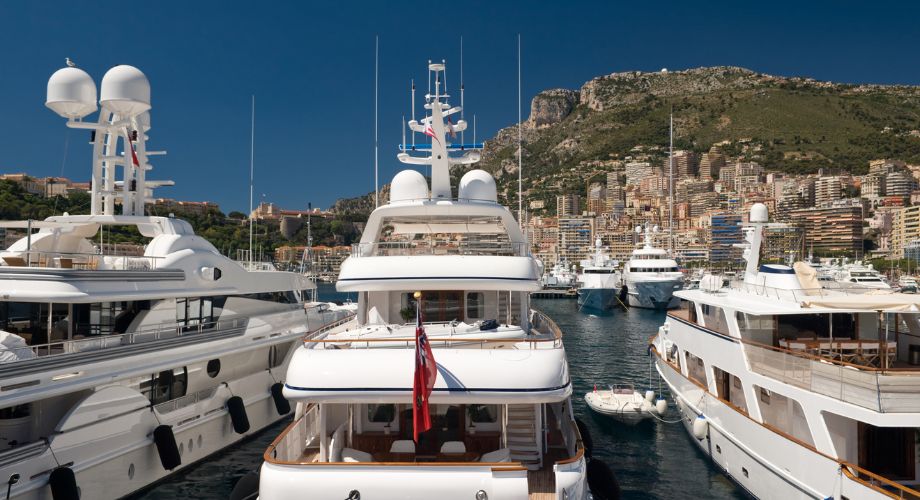 Luxury yachts with various yacht registrations are docked in a scenic marina, set against a hillside cityscape under a clear blue sky.