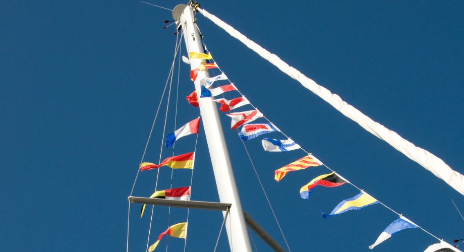 A tall sailboat mast is adorned with a string of vibrant nautical signal yacht flags.