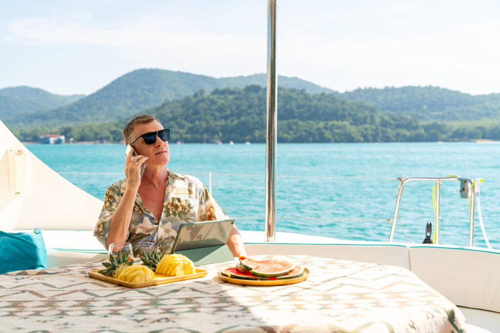 A man in a tropical shirt relaxes on a luxury yacht, enjoying fresh fruit while speaking on the phone, with turquoise waters and lush green hills in the background—a snapshot of the yachting lifestyle.
