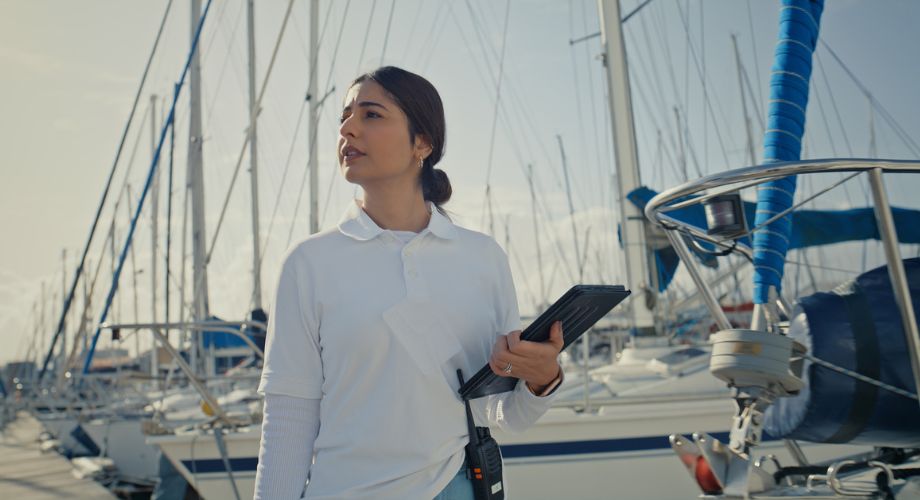 A woman in a white polo shirt holds a tablet and walks through a marina filled with sailboats, appearing to inspect vessels—possibly as part of a boat warranty or yacht warranty evaluation process.
