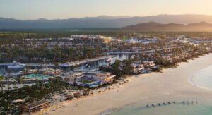 A luxurious coastal resort community featuring beachfront villas, a marina with yachts, lush greenery, and modern architecture set against a backdrop of mountains at sunset.