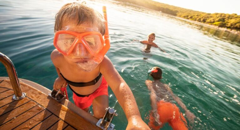 Portrait of boy wearing snorkel standing on steps of luxury yacht with his family swimming in sea in background. Explore the best family cruisers, from small yachts to sailboats, for safe, comfortable, and fun trips at sea.