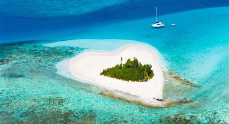 A heart-shaped tropical island surrounded by turquoise waters and coral reefs sits serenely with a white sailboat anchored nearby.