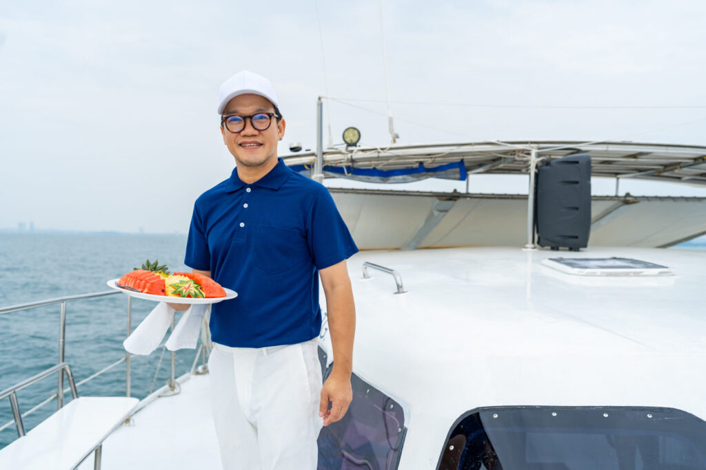 Yacht Cost Breakdown | Buying, Owning & Maintenance | YATCO Portrait of Happy Asian man waiter holding fresh fruit on serving dish for serving to passenger tourist travel on luxury catamaran boat yacht on summer vacation.