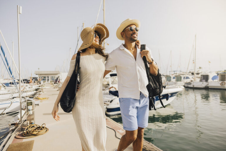 A smiling couple walks hand in hand along the marina's dock, surrounded by boats on a sunny day during their honeymoon yacht charter. Plan the ultimate honeymoon yacht charter. Romantic routes in the Mediterranean, Caribbean, Florida, Greece, Italy, and beyond.