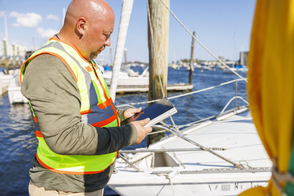 Used Yachts | How to Inspect Before Buying | 2025 Guide | YATCO Mature bald appraiser in safety vest, with digital tablet inspects prow of used yachts in pier in sunny day.