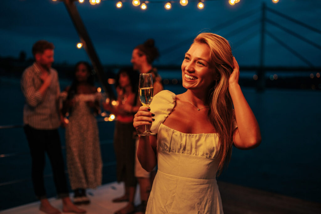 Yacht Entertainment Systems | Tech & Party Features | YATCO A young blonde woman smiles and holds a glass of champagne on a boat ride at night. Discover yacht entertainment systems, luxury options, and tech innovations that elevate charter experiences and unforgettable yacht parties.