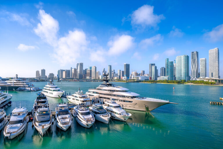 Luxury yachts for sale sit docked on the ocean, set against the Miami skyline and marina.
