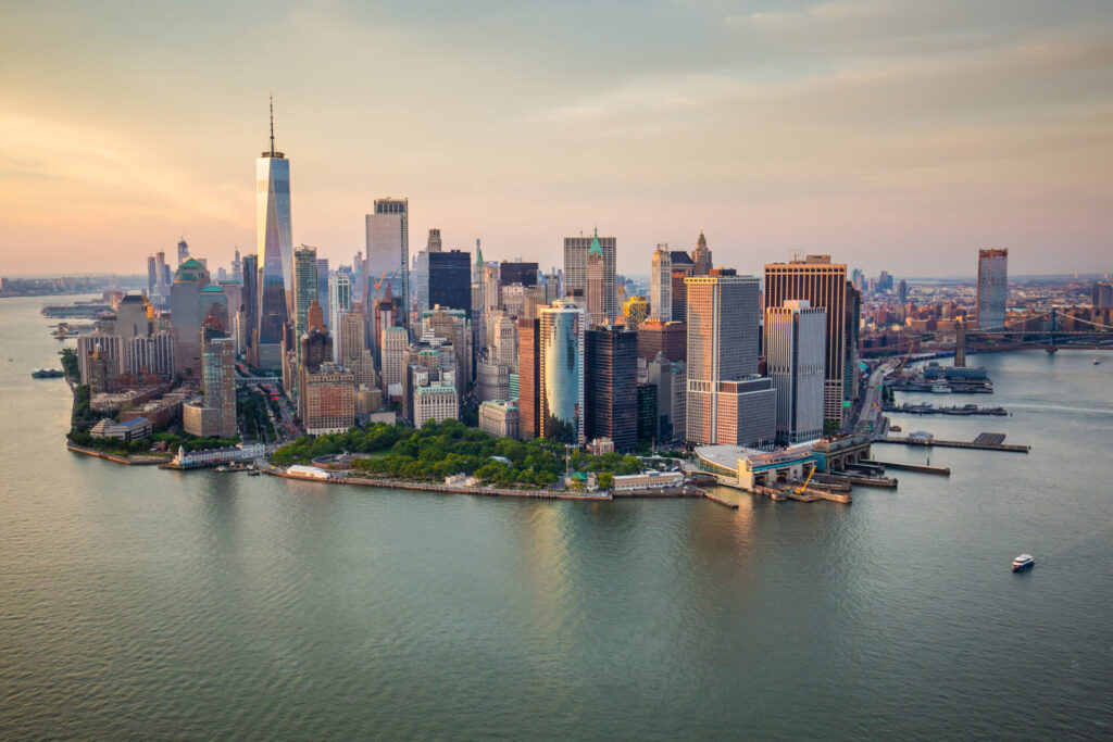 Lower Manhattan at sunset, with warm light illuminating the skyscrapers and the calm waters of the Hudson and East Rivers surrounding the tip of the island.
